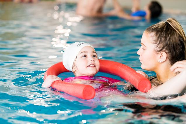 Ein Mädchen mit Schwimmring lächelt in einem Schwimmbecken, während eine Schwimmtrainerin neben ihr steht.